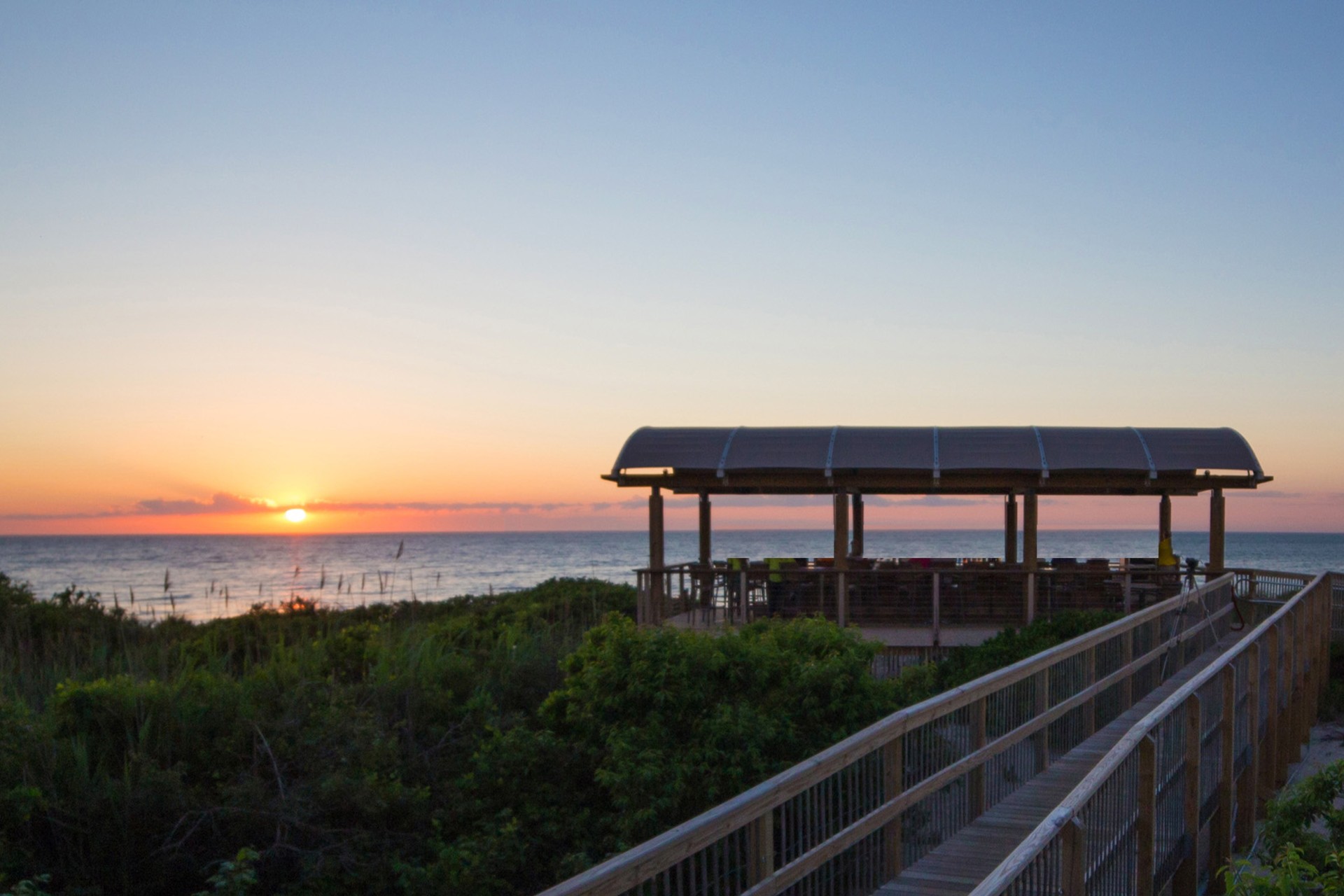 a wooden walkway leading to a beach with a gazebo and bushes