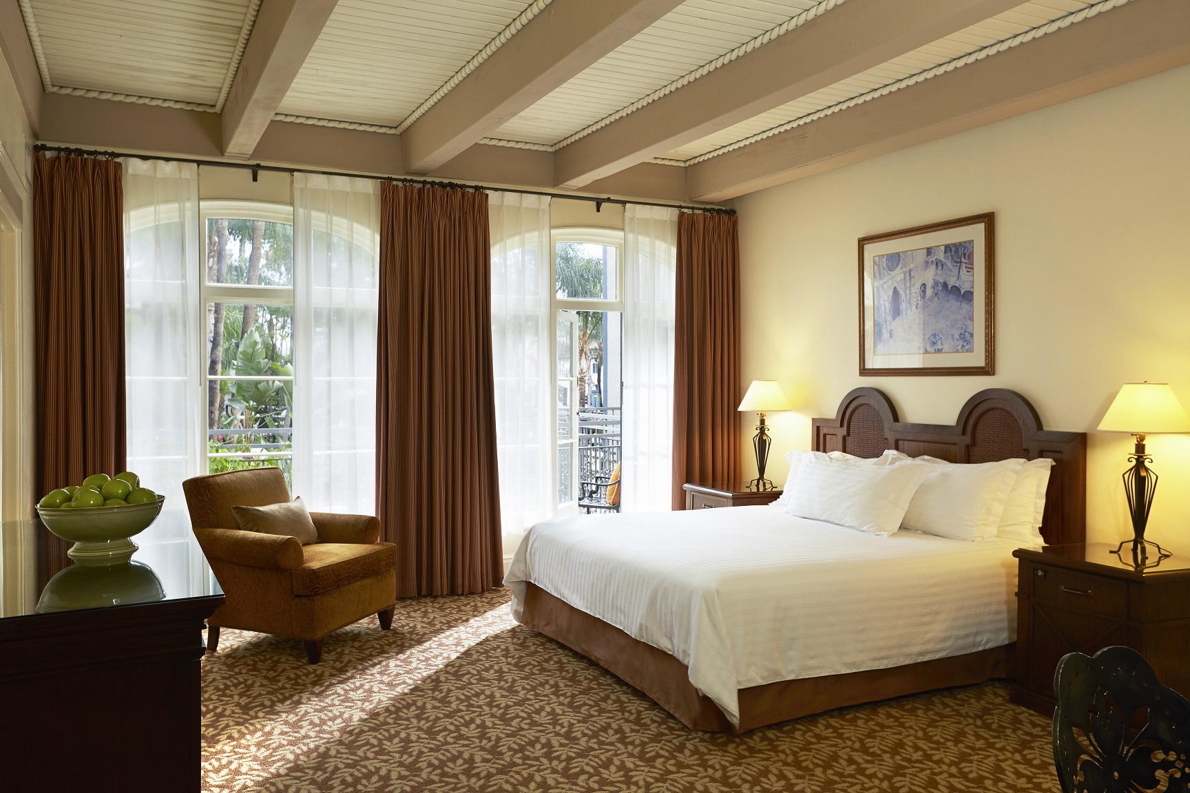 Bright, airy hotel room featuring a king bed with a dark wood headboard, exposed ceiling beams, large French doors with sheer and brown curtains, and an armchair near the window.
