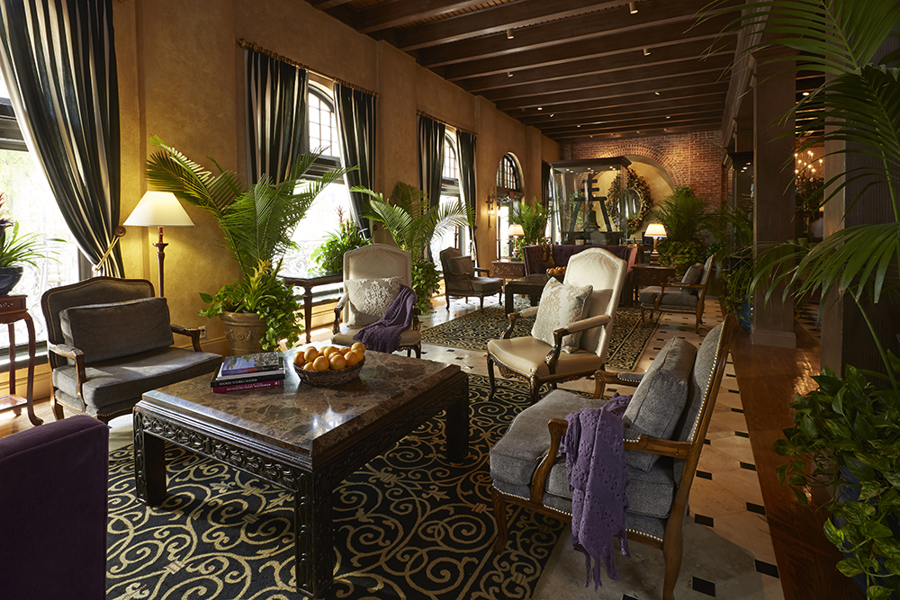 Sophisticated hotel lobby seating area with dark striped drapes, wooden ceiling beams, potted tropical plants, plush armchairs, and carved coffee tables on patterned area rugs.