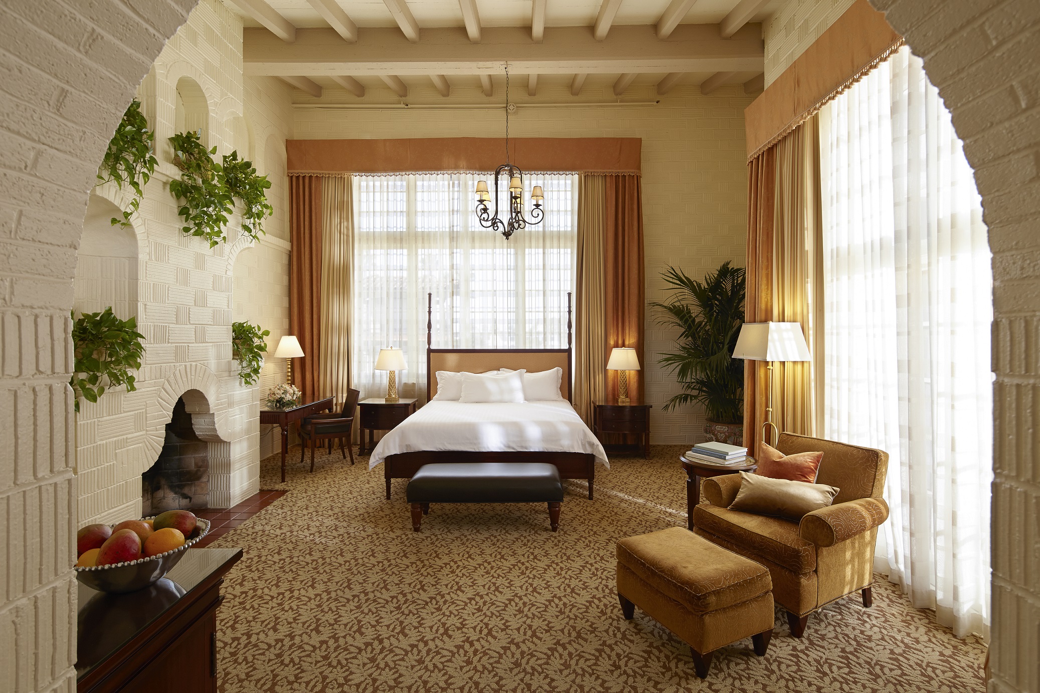 Spacious luxury suite with cream painted-brick arches, a fireplace, high exposed-beam ceilings, four-poster bed, large curtained windows, and a gold-toned armchair with ottoman.