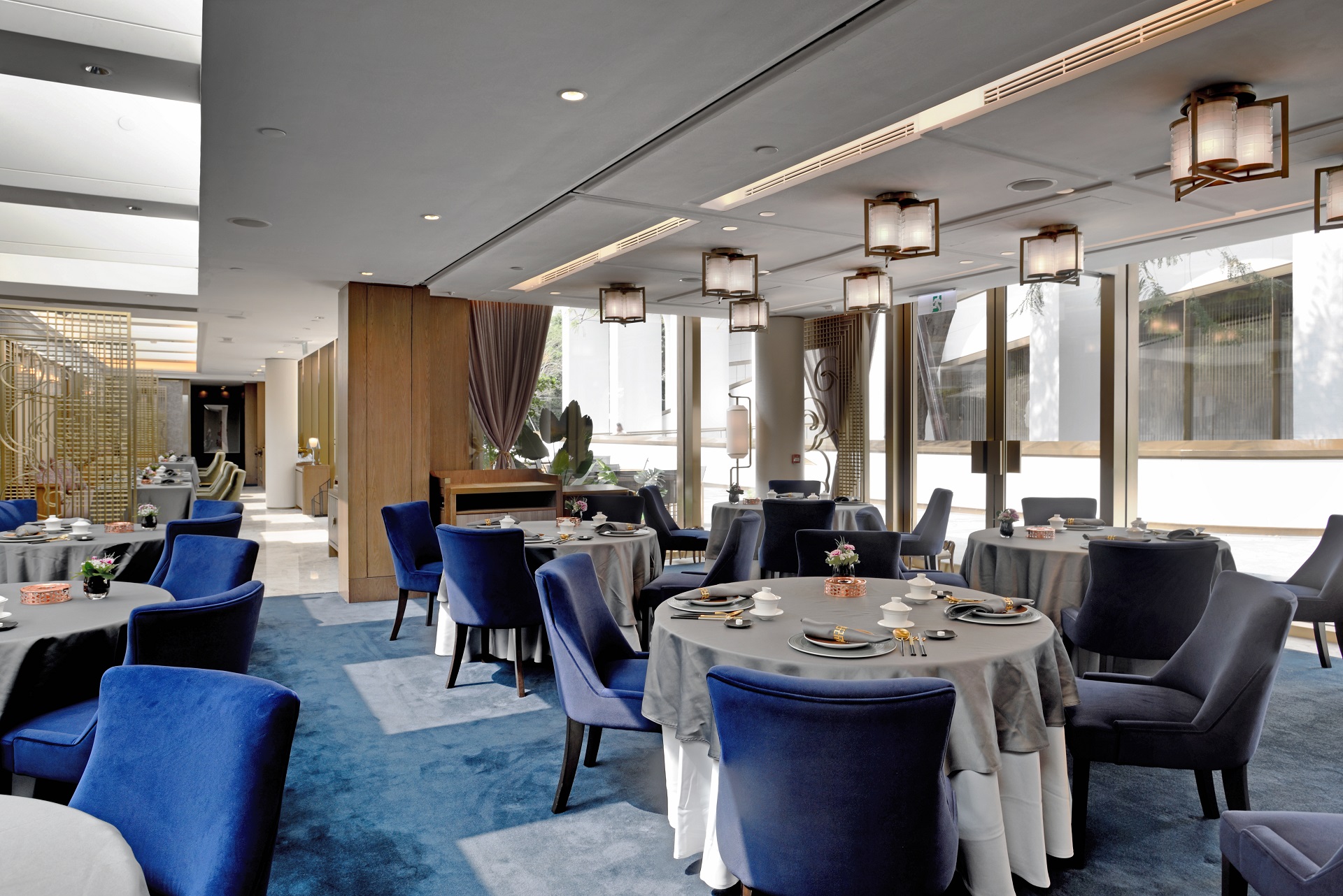 A sophisticated restaurant dining room with blue carpeting, round tables draped in grey and white linens, surrounded by blue velvet armchairs, under a ceiling with contemporary square light fixtures and floor-to-ceiling windows.