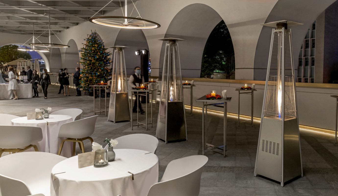 An outdoor terrace set for an evening event with white-draped dining tables, high-top cocktail tables, tall pyramid patio heaters with visible flames, arched stone arcades, and a decorated Christmas tree in the background.