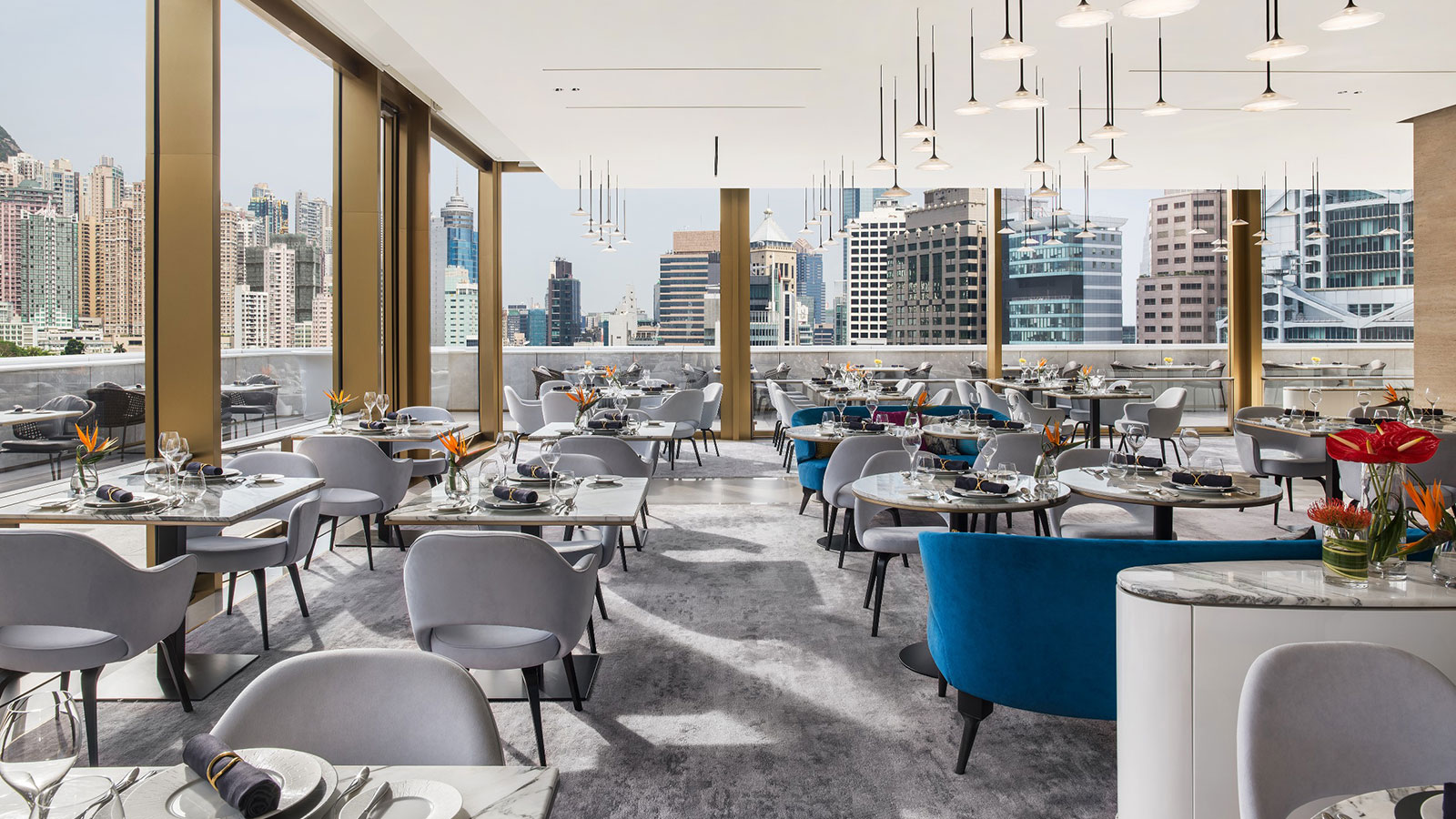 A stylish modern rooftop dining room featuring light grey velvet armchairs, bright blue curved seating, marble-top tables, and delicate pendant lights, with floor-to-ceiling glass windows offering expansive views of a city skyline.