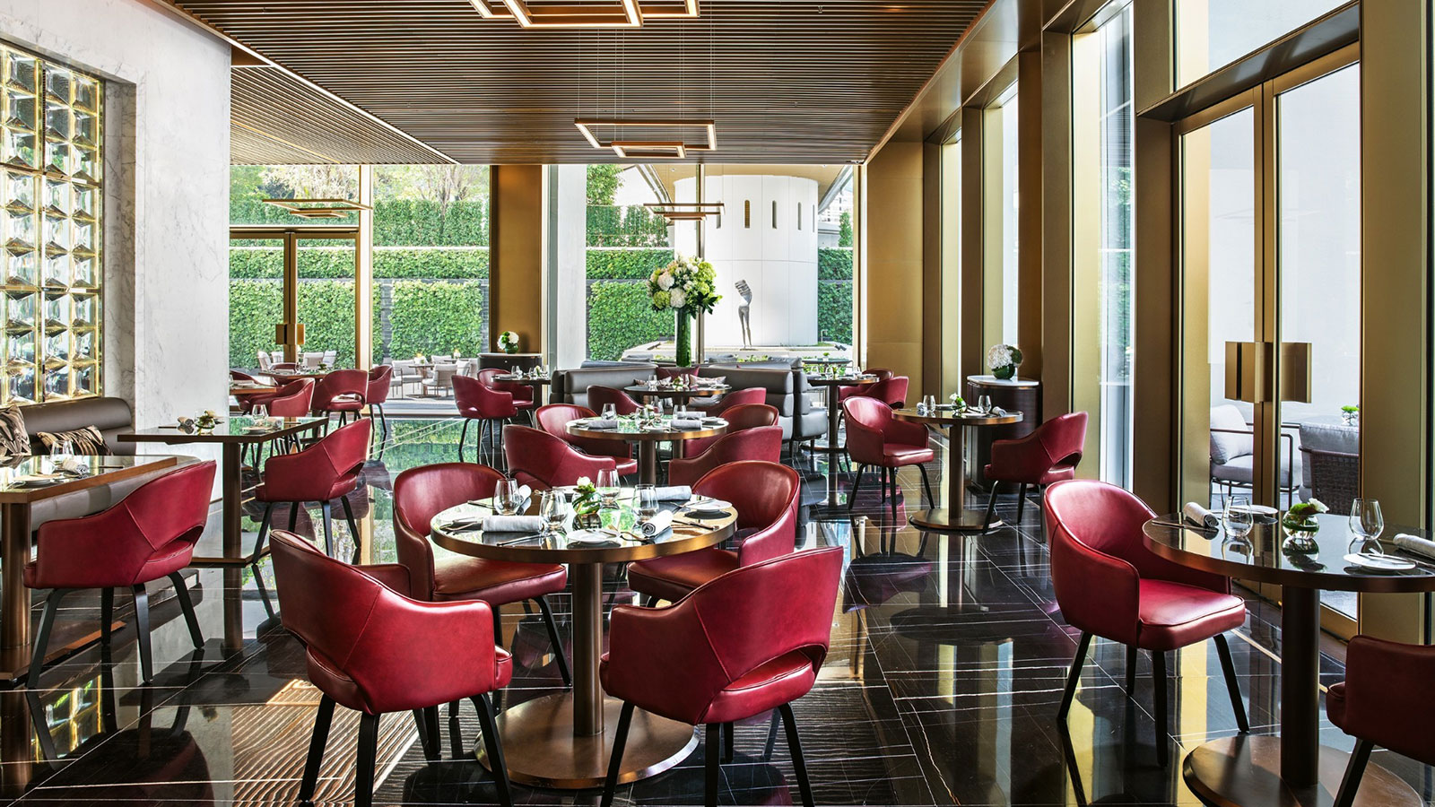 A luxurious, contemporary restaurant dining room featuring polished dark marble floors, round tables with vibrant red leather armchairs, floor-to-ceiling windows overlooking a green outdoor garden wall, and a slatted wood ceiling with modern geometric lig