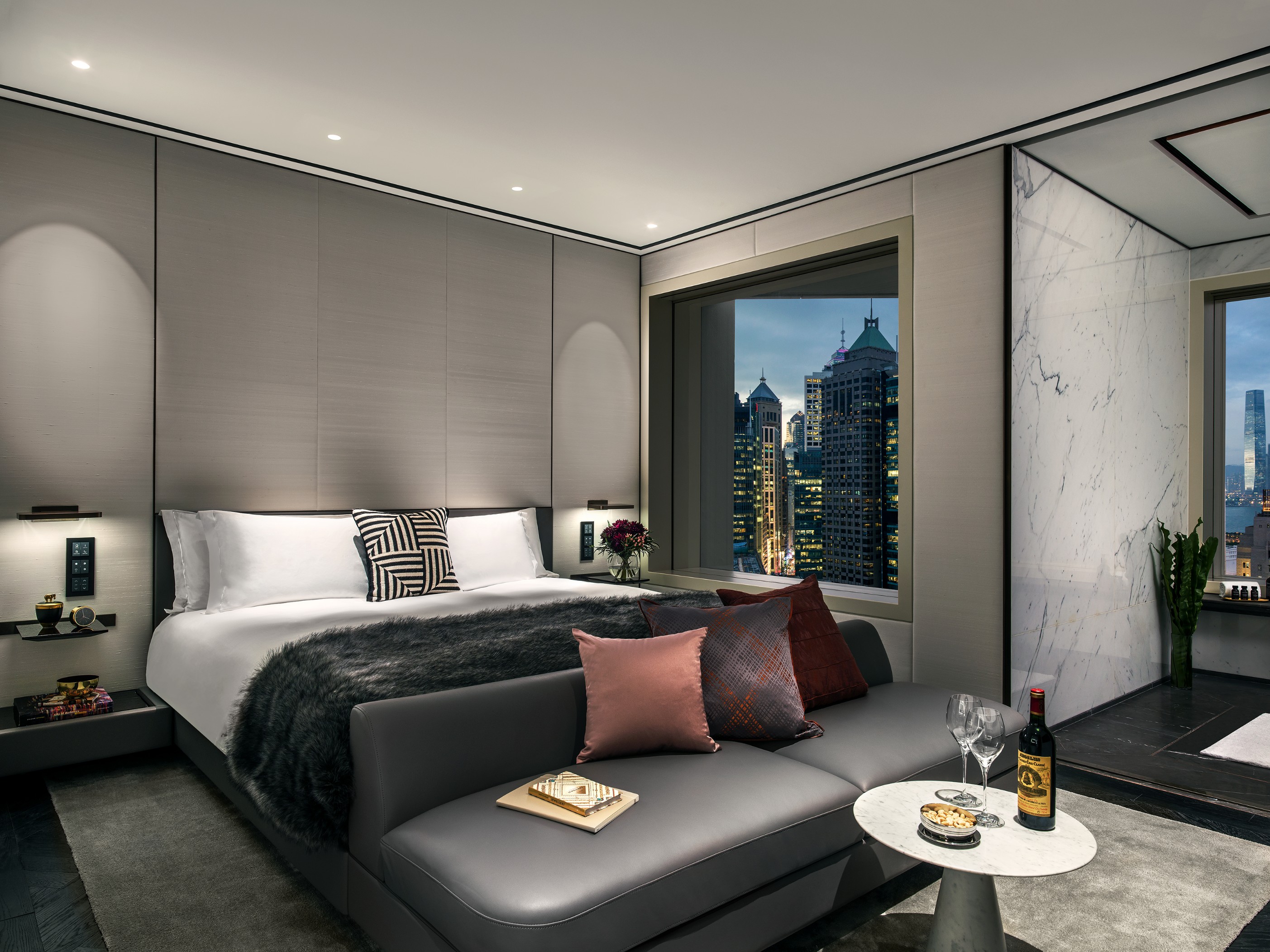 A modern, luxury hotel bedroom featuring a plush bed with grey upholstered paneling behind it, a sleek leather bench at the foot of the bed with decorative throw pillows, a small round coffee table with wine glasses, and a large window framing a illuminat