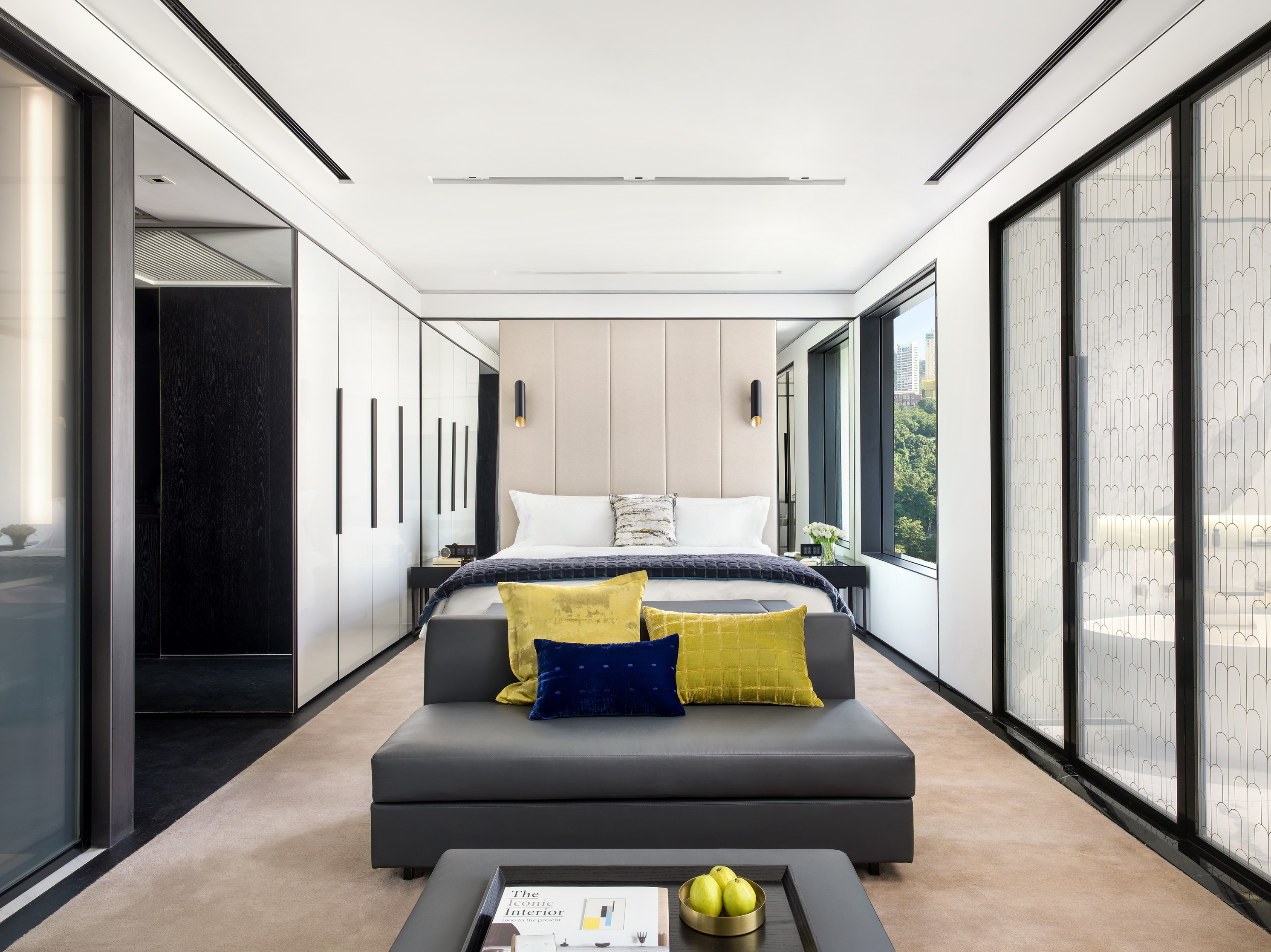 A bright, modern luxury hotel suite with a central king bed backed by a tall upholstered headboard, framed by mirrors and sconces. At the foot of the bed sits a dark grey bench with vibrant yellow and blue accent pillows, leading to a sleek coffee table w