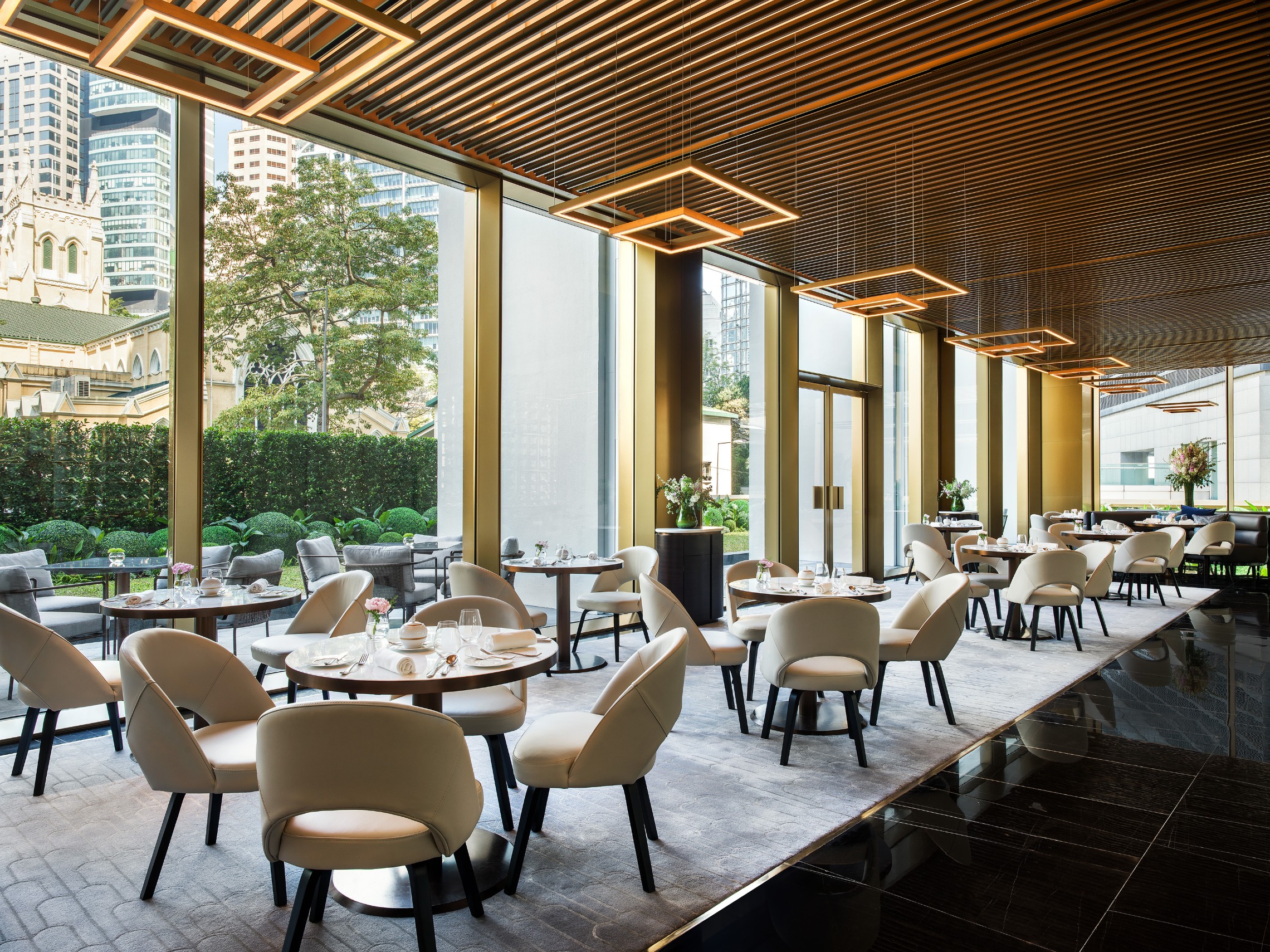 A sophisticated upscale dining room featuring slatted wooden ceilings, geometric square pendant lights, cream armchairs around round wooden tables, and floor-to-ceiling windows looking out onto a garden courtyard and city buildings.