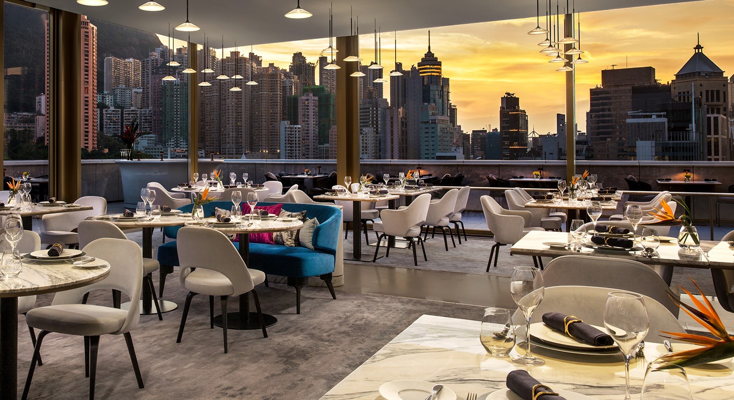 An elegant rooftop restaurant dining room at sunset, featuring sleek white armchairs, a plush teal blue velvet banquette, marble tables set with fine glassware, and large floor-to-ceiling windows framing a golden urban skyline.