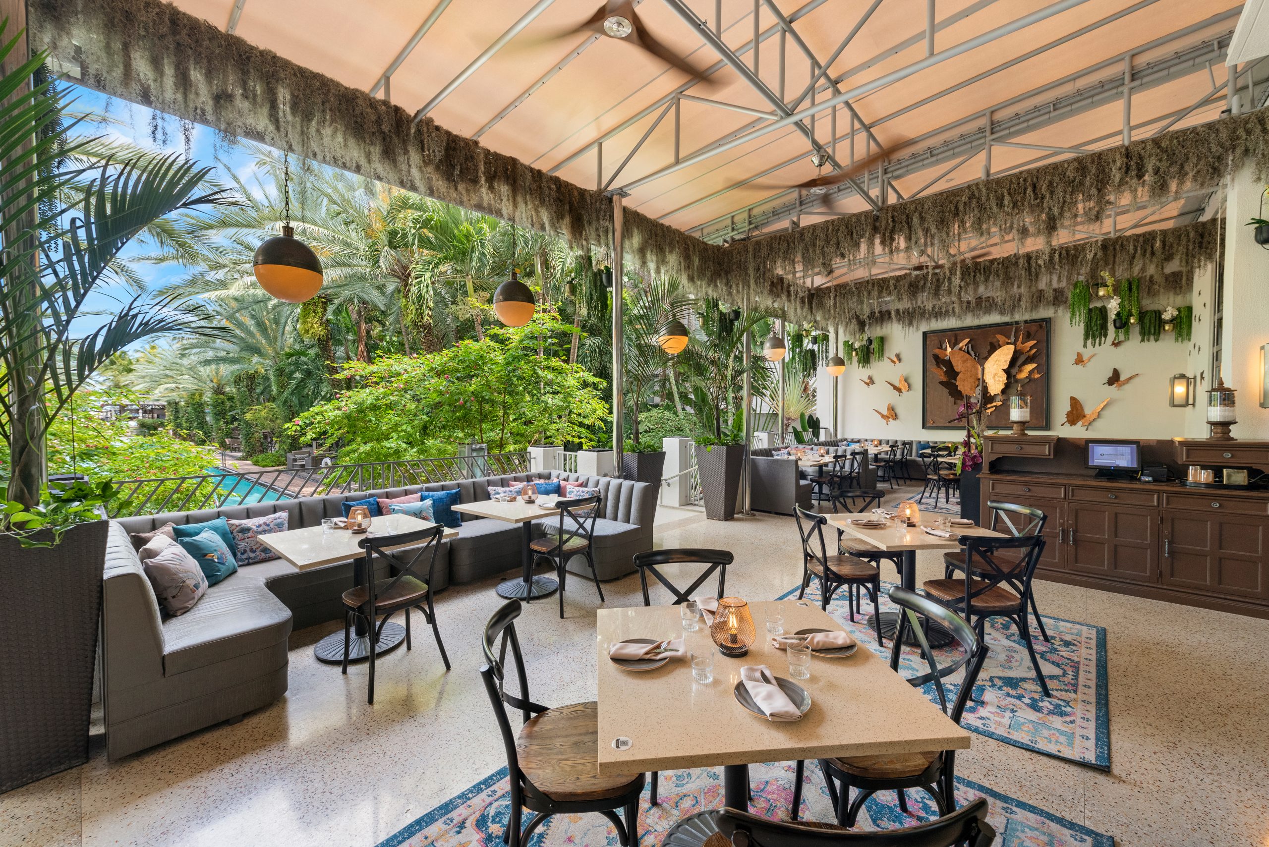 A covered tropical outdoor dining terrace featuring light beige tables, black wooden chairs, plush grey booth seating with colorful cushions, orb pendant lights, and Spanish moss hanging from the ceiling beam.