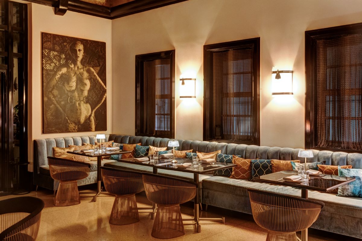 A moody, elegant dining seating area featuring a long plush blue velvet banquet, copper wire chairs, dark tables, wall sconces, and artwork on the wall.