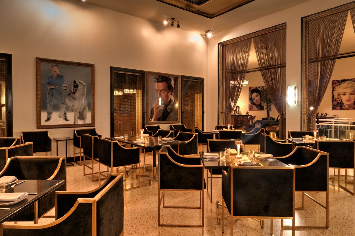 A stylish indoor restaurant dining area featuring black and gold armchairs, dark dining tables with place settings, warm overhead lighting, metallic mesh curtains, and pop art wall portraits.