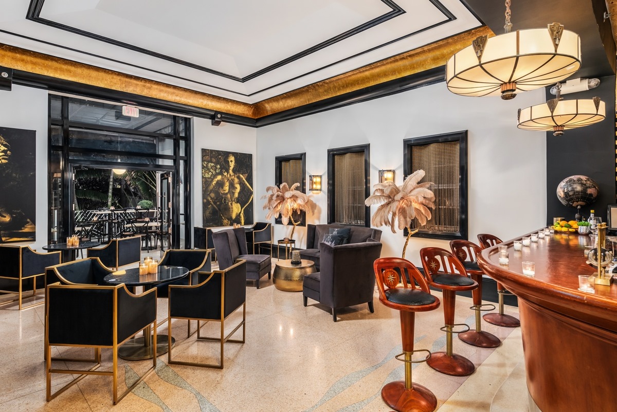 A stylish art deco cocktail lounge featuring a curved wooden bar with stool seating, black velvet armchairs around small tables, feather floor lamps, and gold molding along the ceiling.