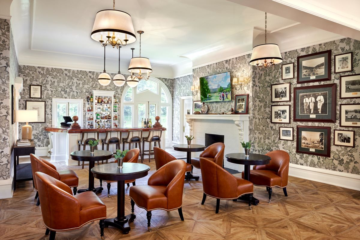 Elegant club lounge featuring floral wallpaper, cognac leather armchairs, small round tables, framed vintage photos, and a bar in the background.