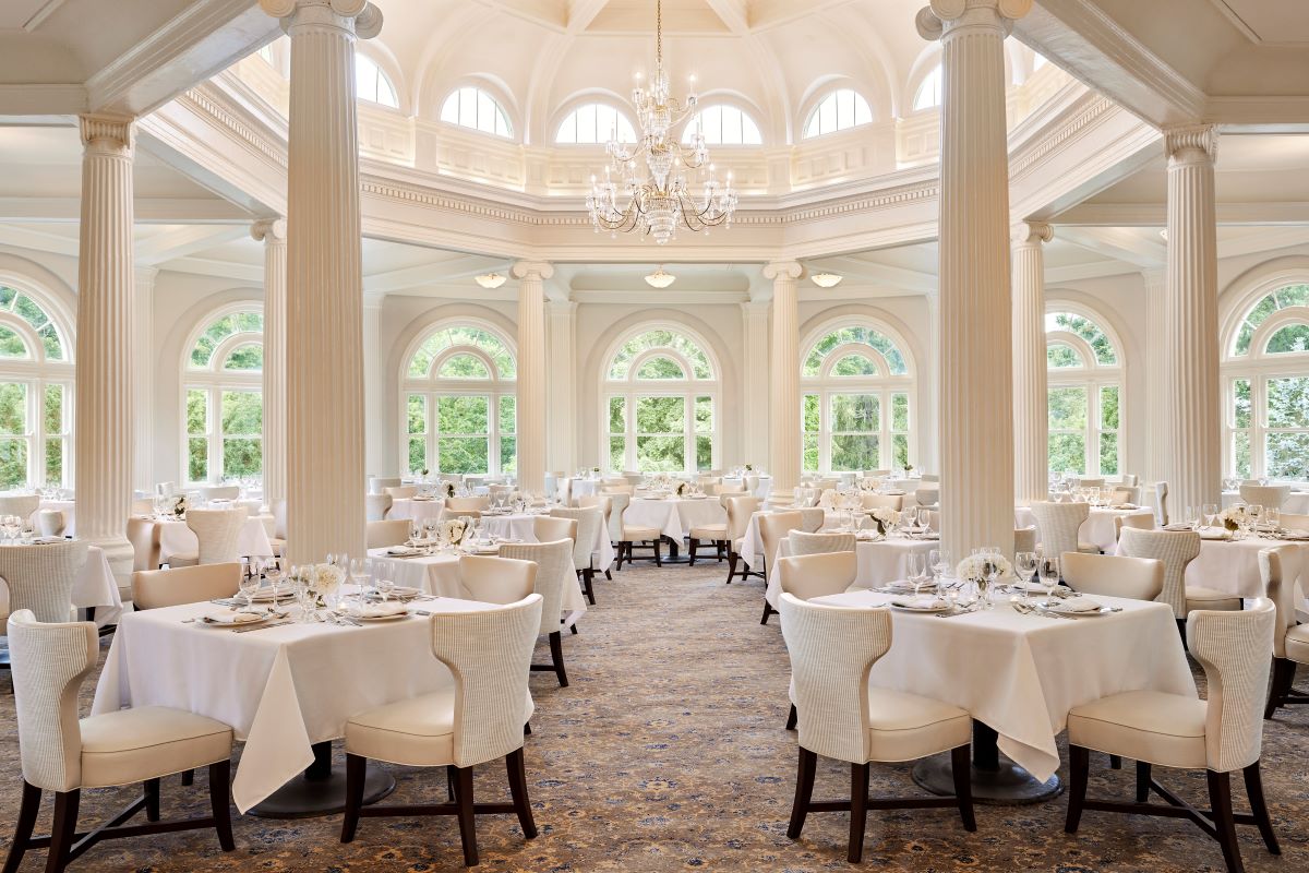 Grand dining room with a central rotunda dome, crystal chandelier, white columns, arched windows, and elegant tables set with white tablecloths.