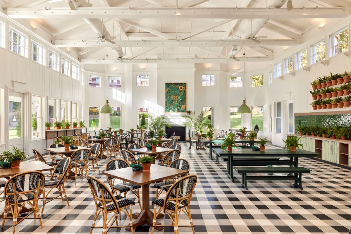 Bright and airy garden house dining space with checkered flooring, high white vaulted ceiling, woven bistros chairs, green picnic tables, and potted plants throughout.