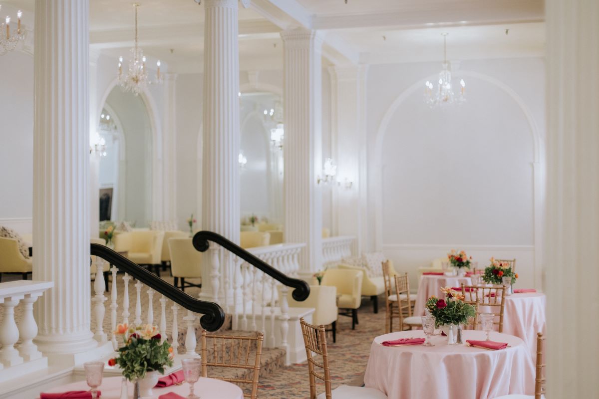 Elegant interior event space featuring classical white columns, balustrades, crystal chandeliers, and dining tables decorated with light pink tablecloths, floral centerpieces, and gold Chiavari chairs.