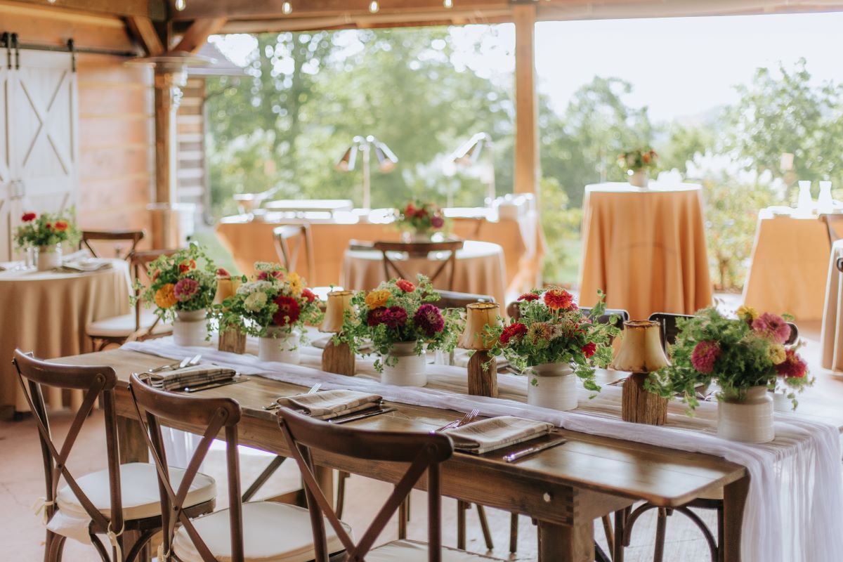 Rustic outdoor wedding setup with wooden tables, vibrant floral centerpieces, small lamps, and covered pavilion seating.