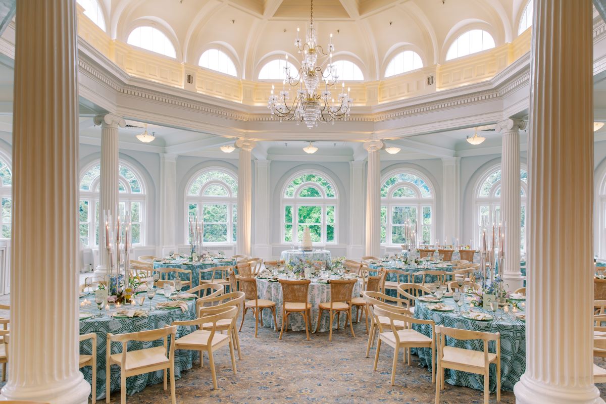 Grand rotunda ballroom set for a wedding reception with patterned light blue tablecloths, modern light wood chairs, tall candelabra centerpieces, and a central crystal chandelier under a domed ceiling.