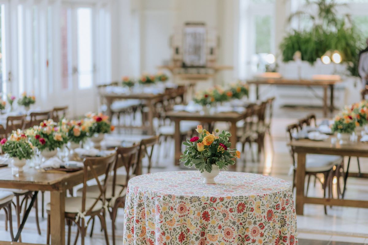 Bright venue decorated for an event with wooden dining tables and a round cocktail table draped in a colorful floral tablecloth, featuring colorful flower centerpieces.