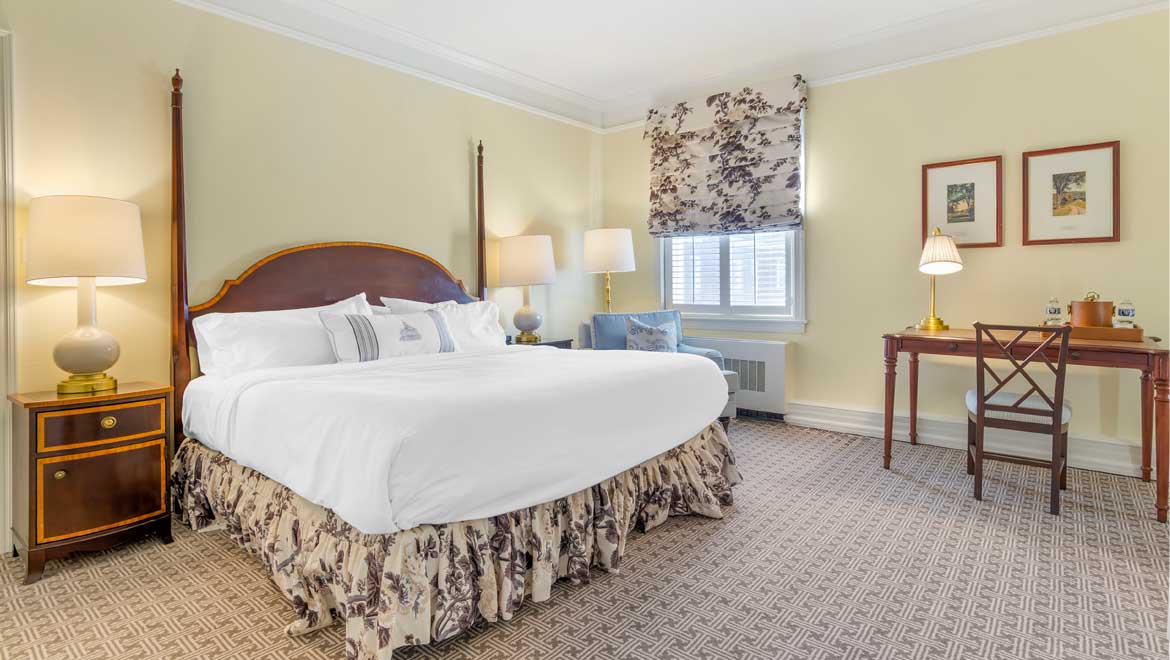 Bright hotel bedroom with pale yellow walls, a four-poster king bed with a floral dust ruffle, and a wooden writing desk.