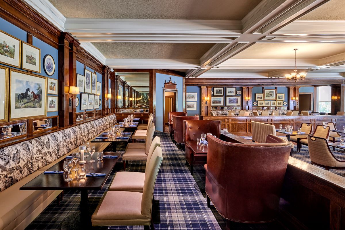 Elegantly styled restaurant dining room featuring blue walls with framed artwork, dark wood paneling, plaid carpet, and leather armchair seating.