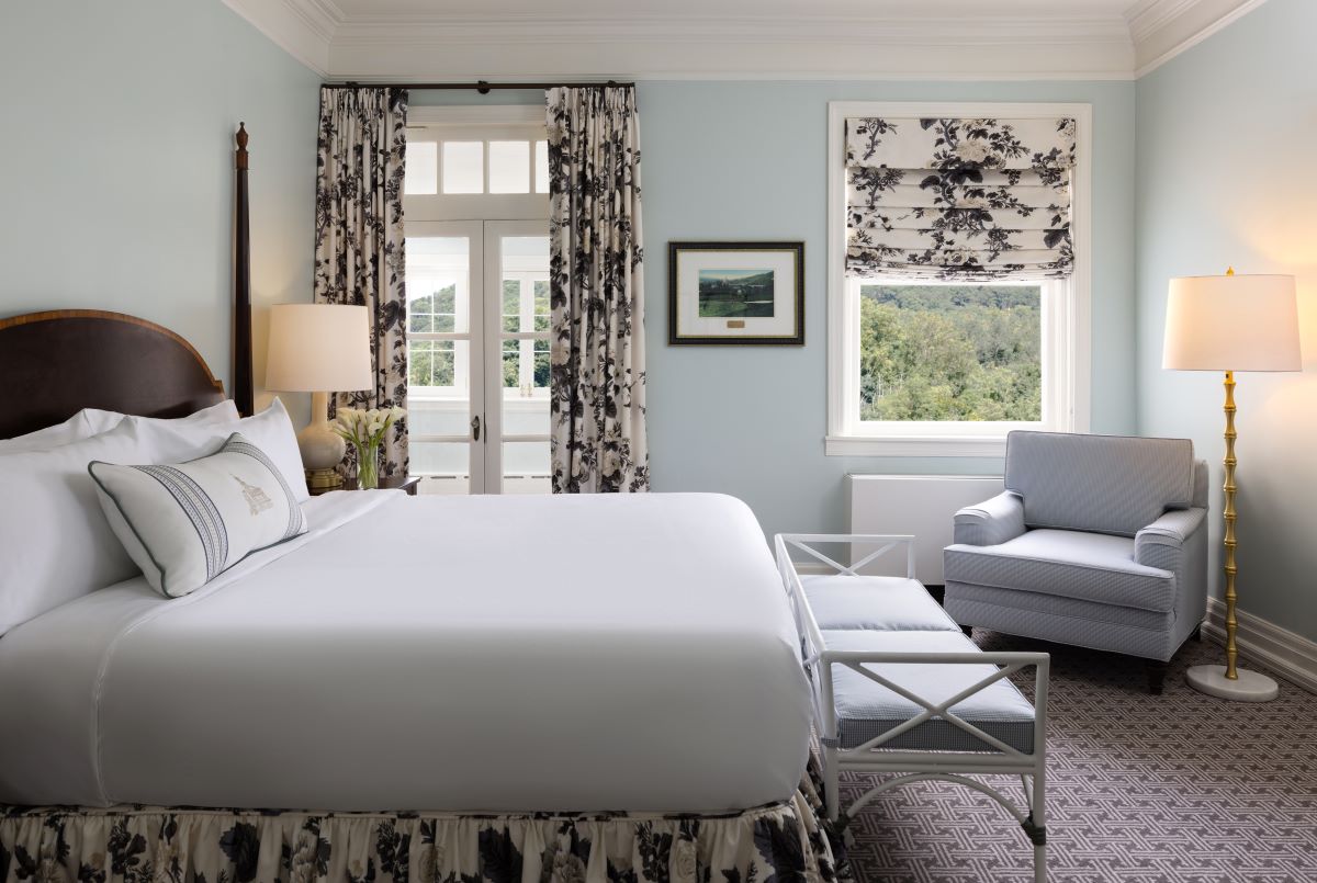 Elegant hotel bedroom with pale blue walls, a four-poster bed made with crisp white linens, floral curtains, a matching floral bed skirt, a light blue armchair, and French doors leading out to a balcony.