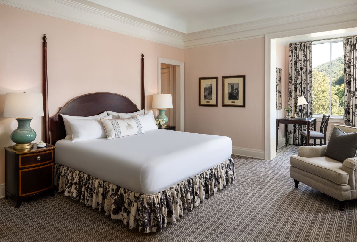 Elegant hotel room with light pink walls, four-poster wooden bed, dark wood nightstands, and a sunlit alcove seating area.