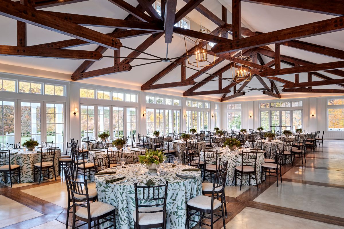 Spacious pavilion reception hall with vaulted dark timber roof trusses, modern geometric chandeliers, floor-to-ceiling glass doors, and round tables with leaf-patterned tablecloths and black Chiavari chairs.