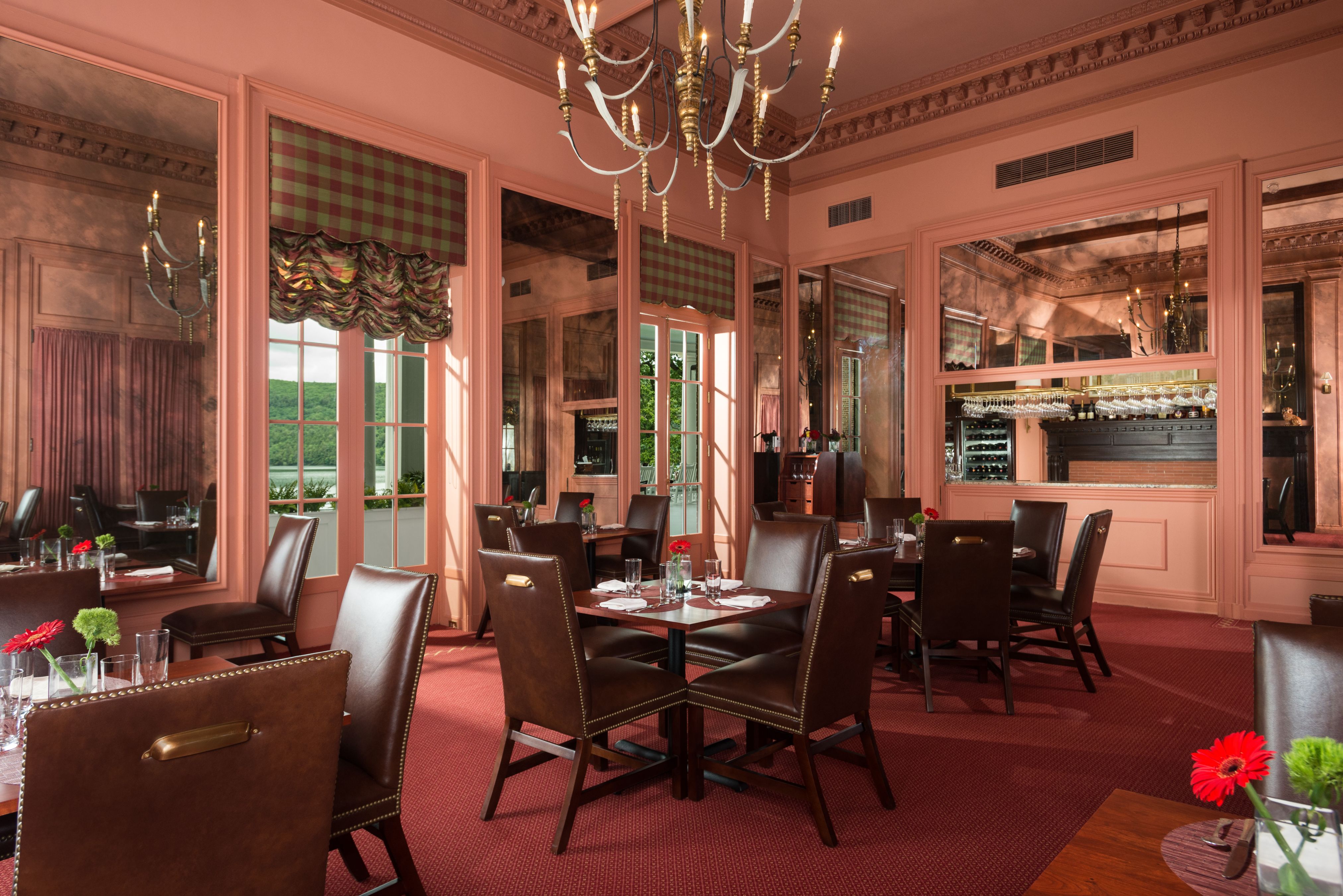An elegant dining room featuring warm salmon-pink walls, large wall mirrors, dark brown leather dining chairs around dark wooden tables, red carpeting, and a classic metal chandelier.