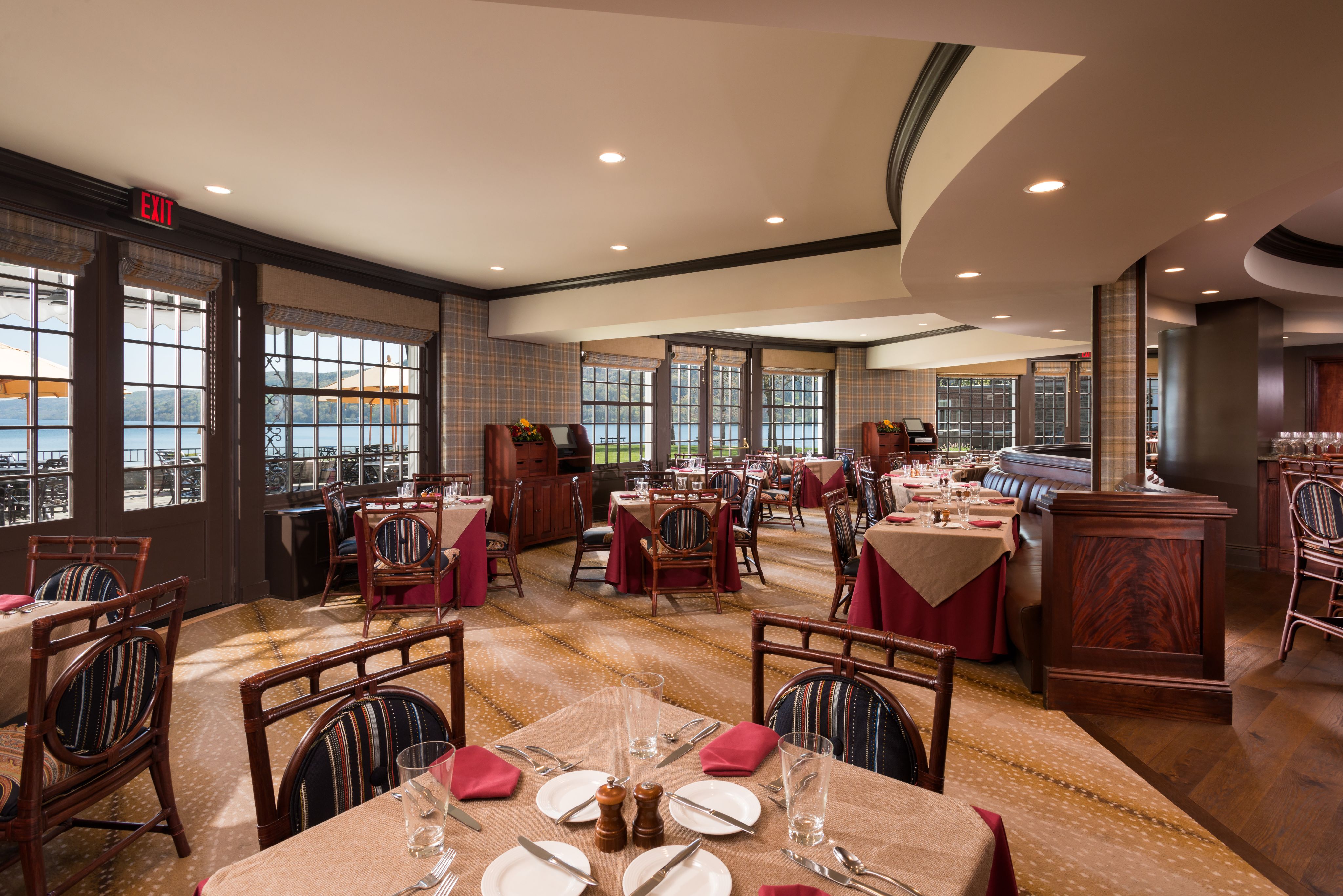 A spacious resort dining area with large windows offering views of a lake, featuring brown wooden chairs with striped cushions, tables set with beige linens and red napkins, and warm interior lighting.