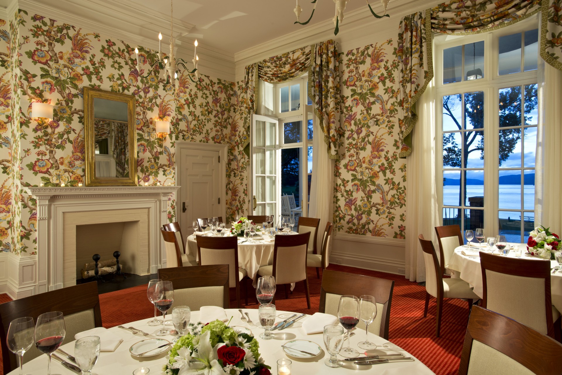 An elegant dining room featuring vibrant floral wallpaper, a classic white fireplace with a gold mirror above, round tables with white tablecloths and wine glasses, and large windows revealing a scenic lake view at dusk.