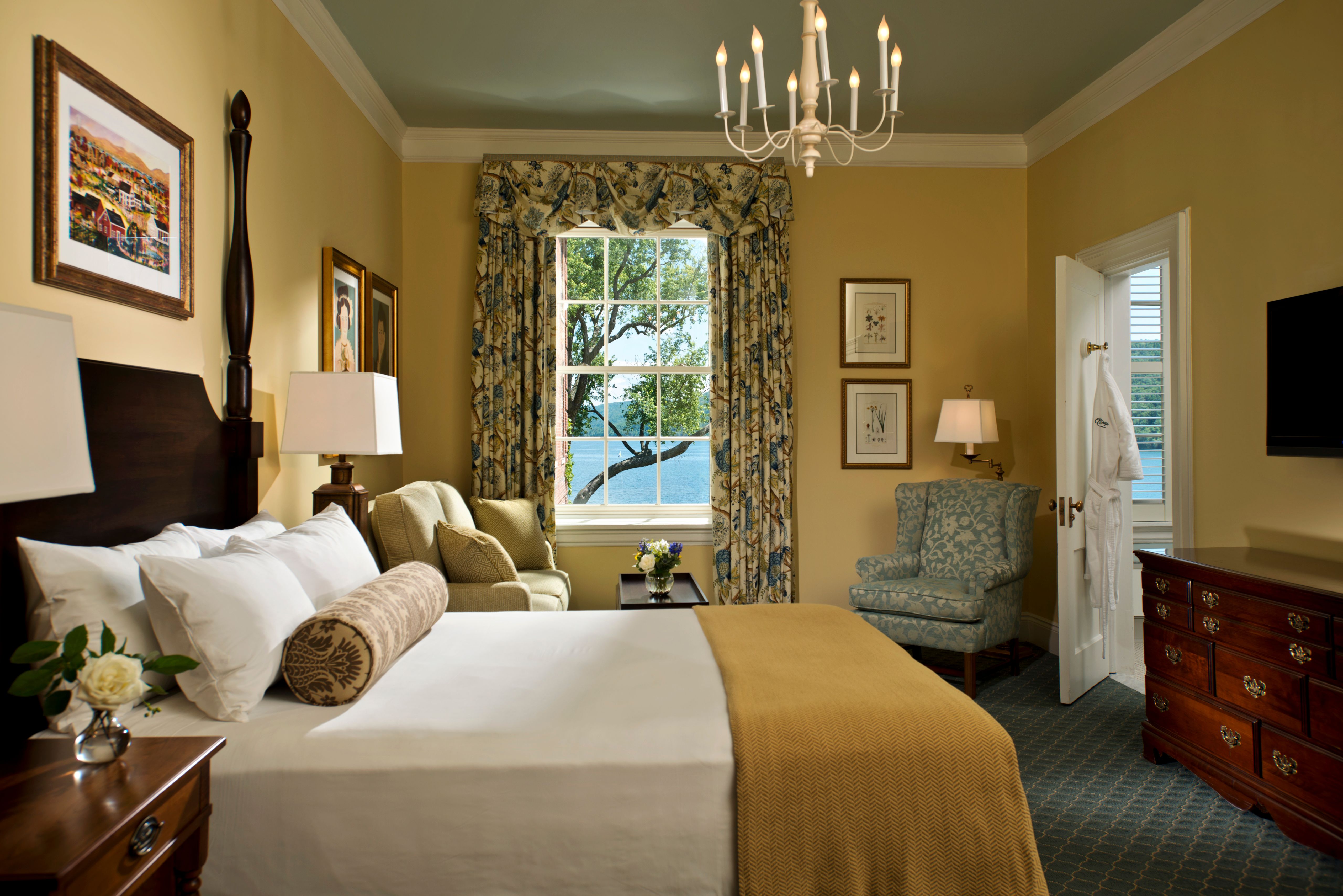 A warm, elegantly decorated hotel bedroom featuring a dark wood bed with white linens and a gold throw, a window overlooking a body of water, a floral armchair, and traditional furniture.