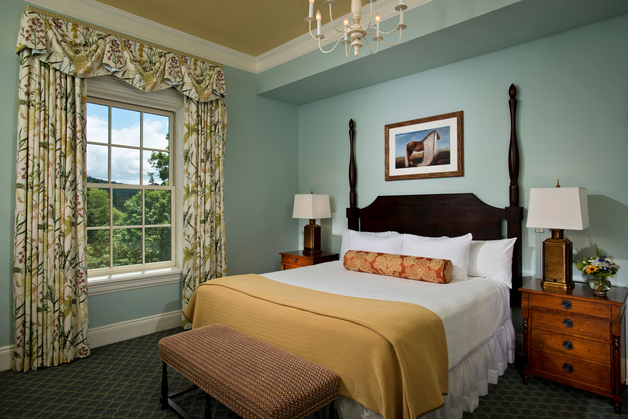 A stylish guest bedroom with pale blue walls, a dark wood post bed dressed in white linens and a mustard yellow blanket, a window with botanical print drapes overlooking greenery, and a chandelier overhead.