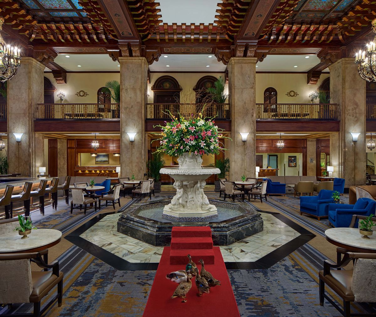 Grand hotel lobby featuring an ornate wooden ceiling, marble columns, a central carved fountain with a large floral arrangement, a red carpet runner in the foreground with live ducks, and plush seating throughout.