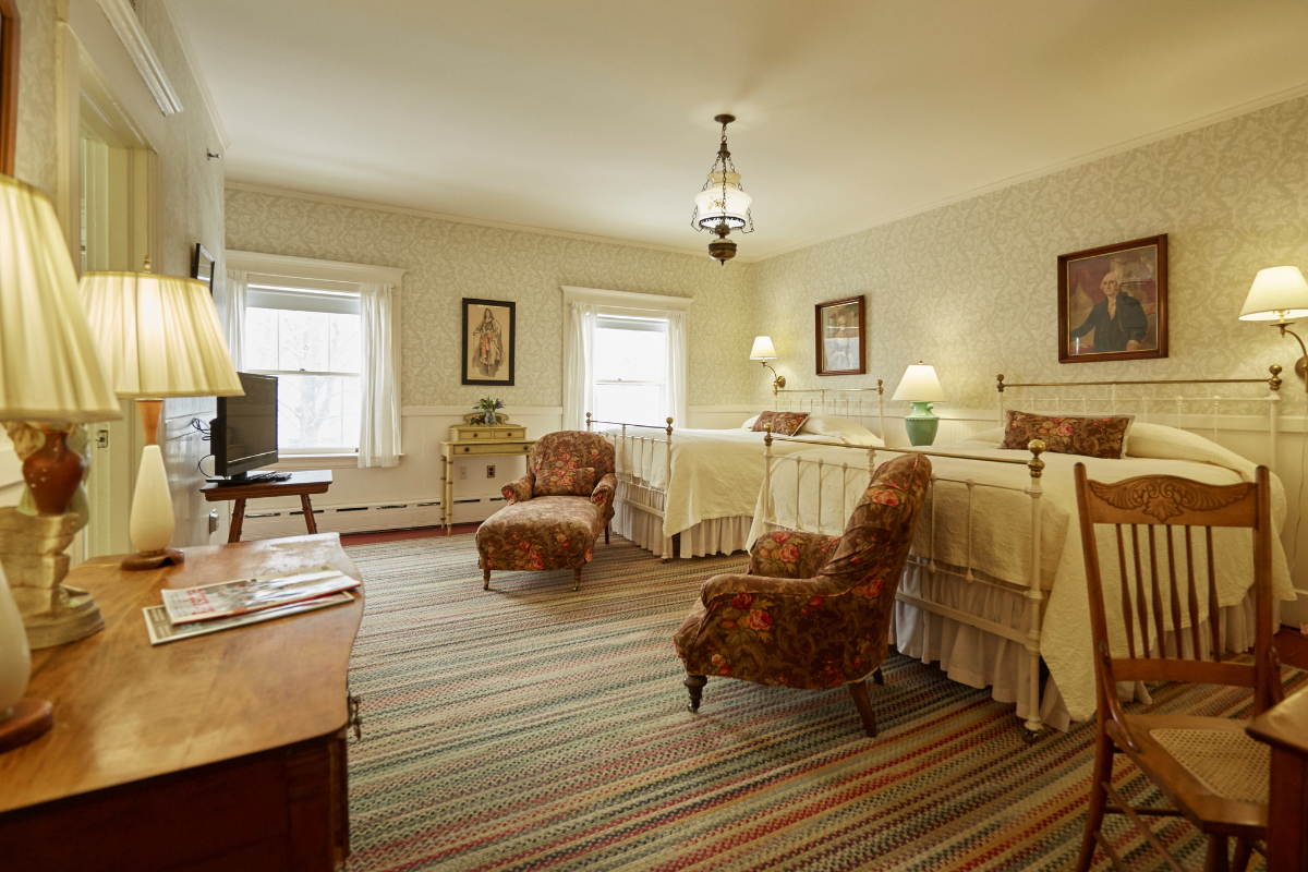 Spacious vintage-style bedroom with two white metal beds, floral armchairs, damask patterned wallpaper, and a striped area rug over dark flooring.