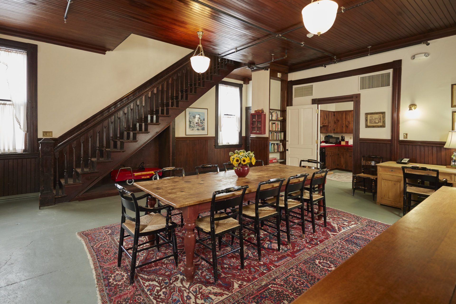 Rustic dining room featuring a long wooden table with black woven chairs, a red patterned rug, dark wood ceiling paneling, and an open wooden staircase along the wall.