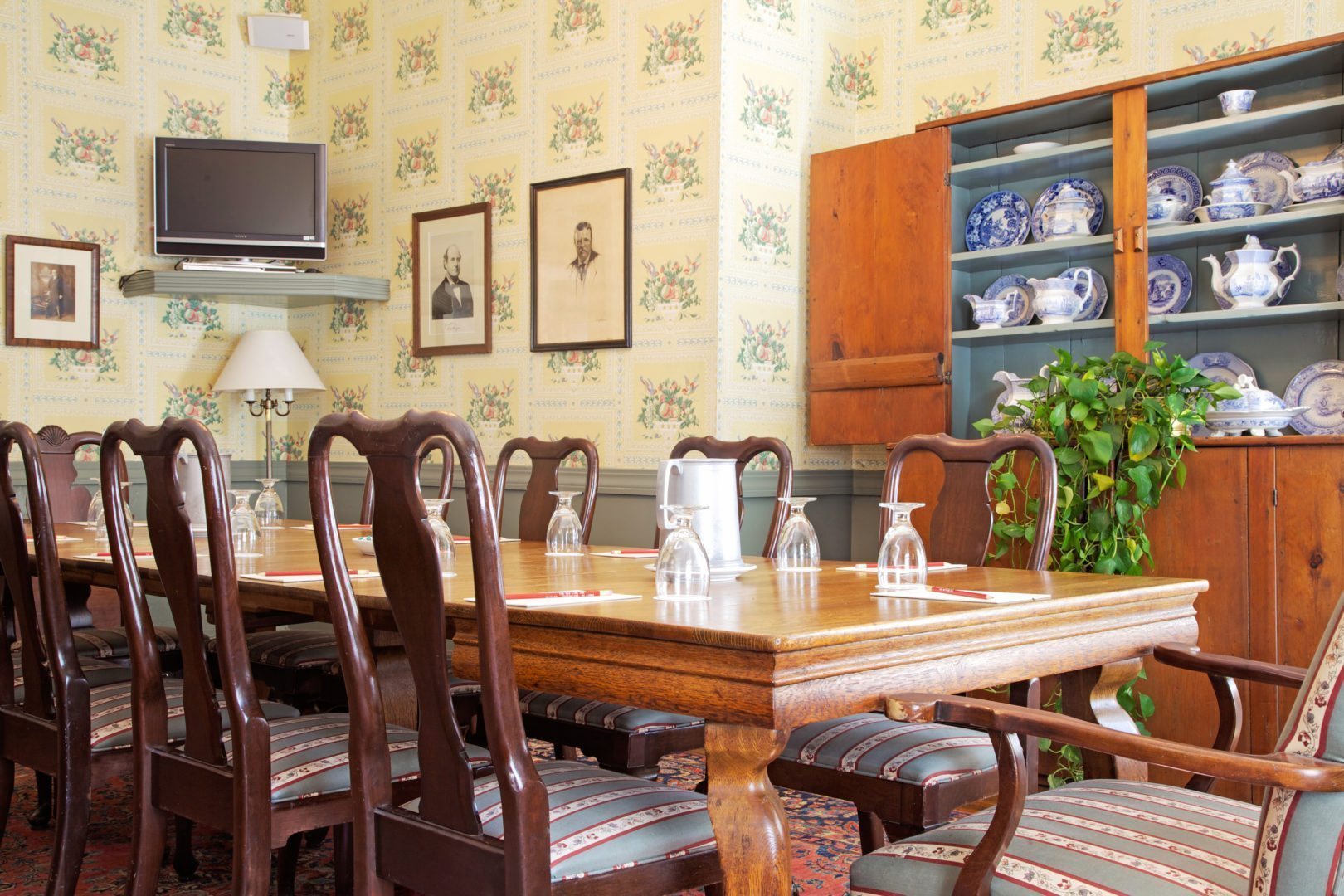 Classic dining or meeting room featuring a long wooden table set with glasses, floral wallpaper, framed historic portraits, and a large wooden cabinet displaying blue-and-white china.