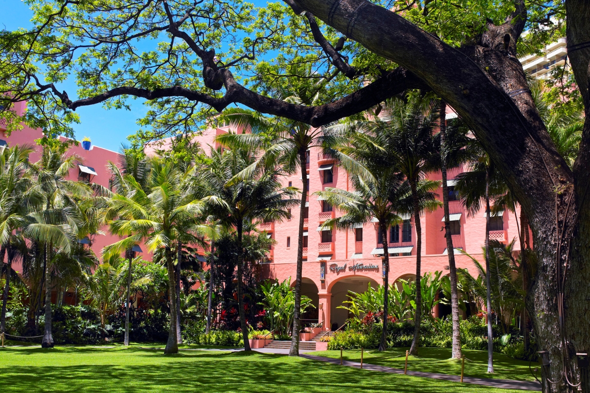 A bright exterior view of the iconic pink facade of The Royal Hawaiian resort, framed by lush tropical palm trees, a large shady banyan tree, and a green lawn.