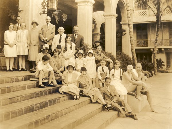 Vintage sepia-toned photograph of a large group of men, women, and children gathered together outdoors on wide steps in front of a Spanish-style archway and courtyard building.