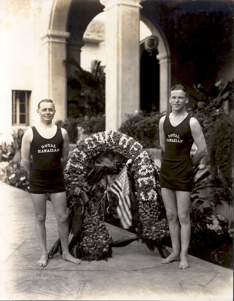 two men in swimsuits standing next to a flower arch