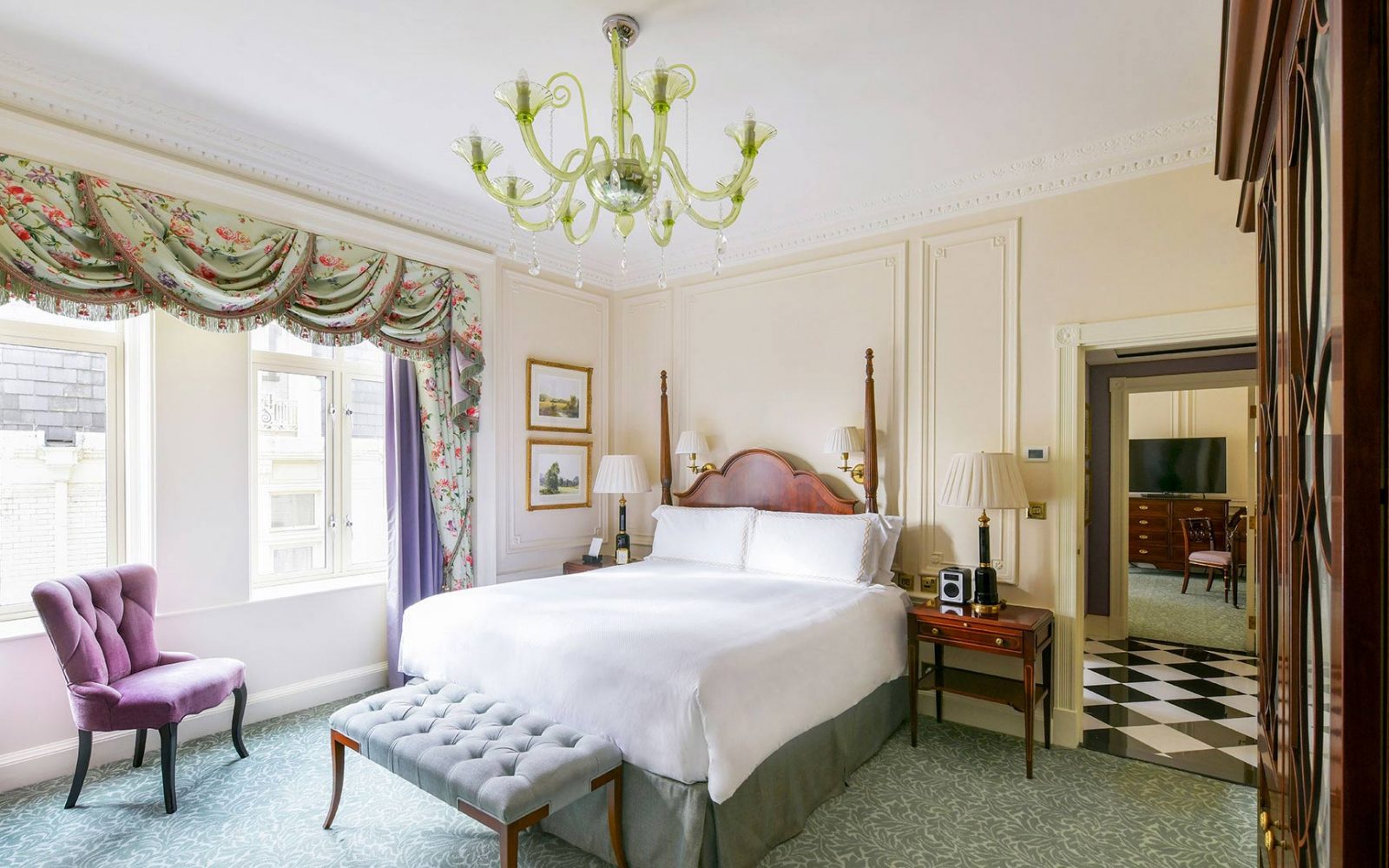 Elegantly decorated hotel suite bedroom featuring a wooden four-poster bed with crisp white linens, a lime green glass chandelier, floral window drapery with a swaddled valance, a purple tufted armchair, and an adjacent entryway with black-and-white check