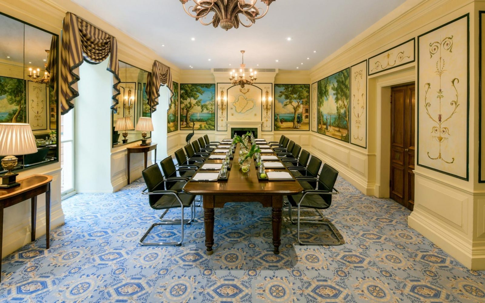 A grand boardroom featuring a long wooden conference table with black leather armchairs, intricate painted wall murals, a blue patterned carpet, chandeliers, and mirrored wall panels.