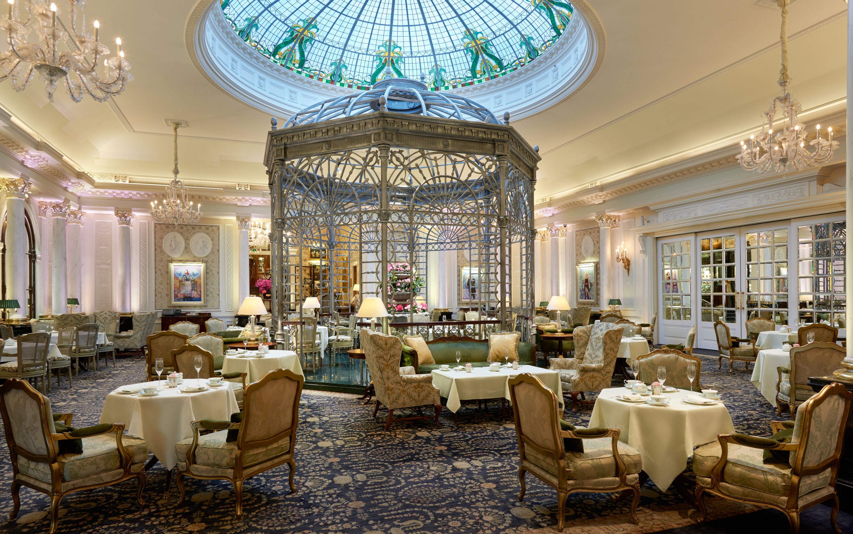 A grand, opulent dining salon featuring an ornate metal birdcage gazebo structure under a circular stained-glass skylight, surrounded by white-clothed tea tables, plush armchairs, and crystal chandeliers.