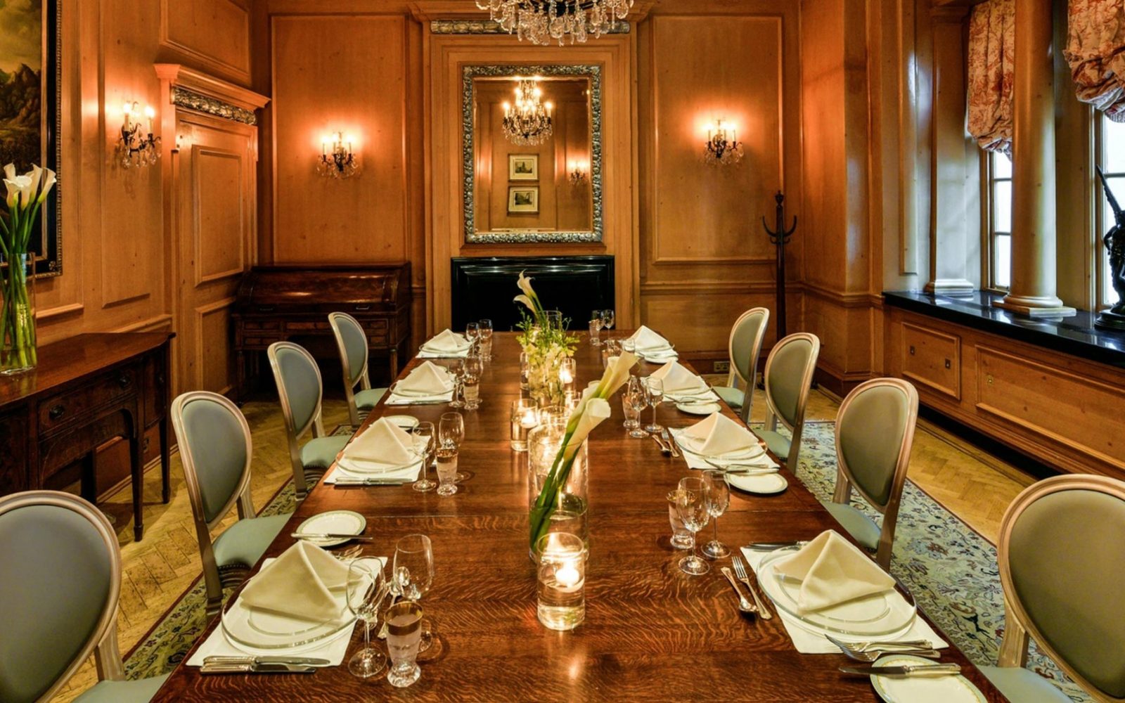 A private dining room featuring warm wood-paneled walls, a long polished wooden dining table set for a multi-course meal, grey upholstered oval-backed chairs, glowing candles, white calla lily centerpieces, and a crystal chandelier reflected in a ornate m