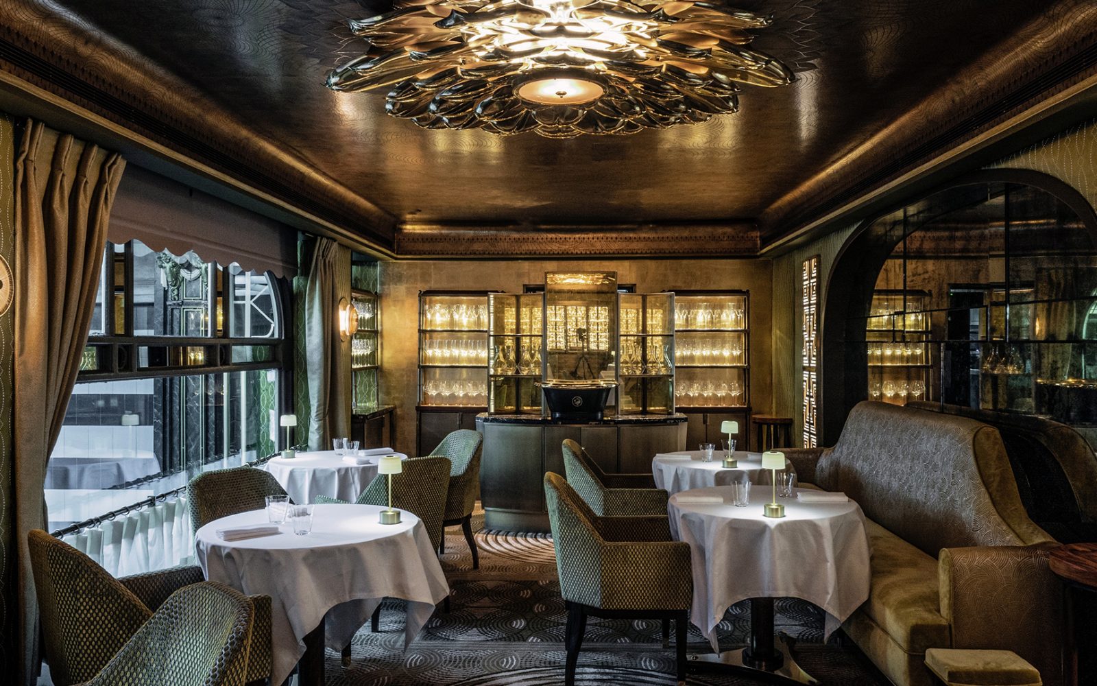 A moody, sophisticated dining area featuring low warm lighting, an ornate metallic ceiling centerpiece, white-draped tables, green textured armchairs, a plush banquette, and illuminated glassware display cabinets in the background.