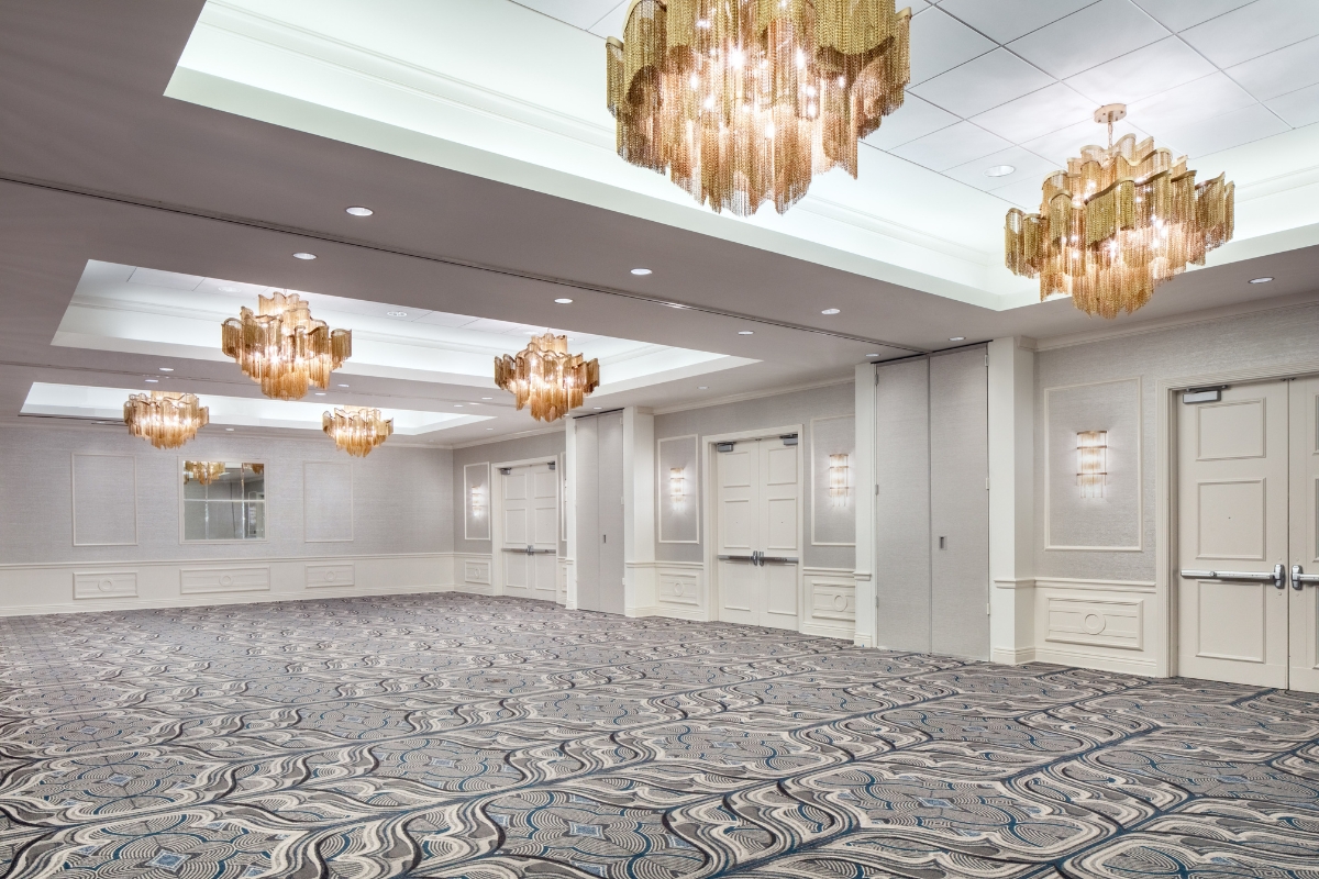 Large empty ballroom with patterned blue and grey carpet, light grey walls with white wainscoting, multiple white double doors, wall sconces, and tiered gold fringe chandeliers hanging from recessed ceiling bays.