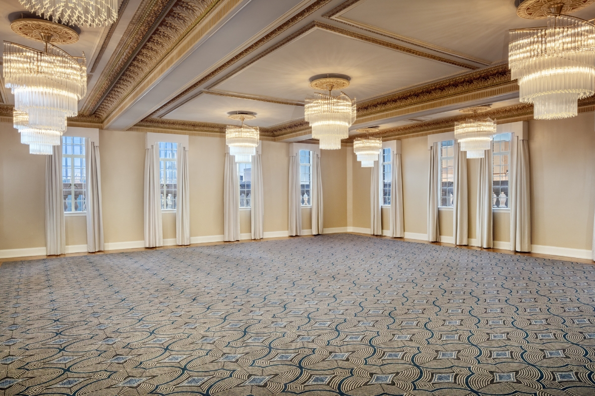 Spacious empty ballroom with elaborate gold ceiling molding, several large tiered crystal chandeliers, patterned grey and blue carpet, neutral beige walls, and tall windows framed with cream drapes.