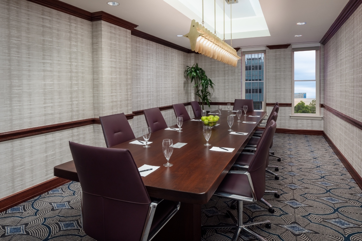 Executive boardroom featuring a long dark wood conference table lined with deep purple leather armchairs, place settings with writing pads and water glasses, a central bowl of green apples, a modern multi-pendant light fixture hanging from a recessed ceil