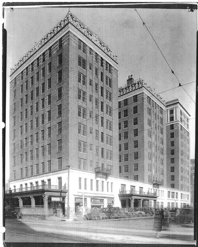 Vintage black-and-white photograph showing a tall, historic brick hotel building with ornate top-ledge detailing, street-level storefronts including a drug store, and early 20th-century automobiles parked along the street outside.