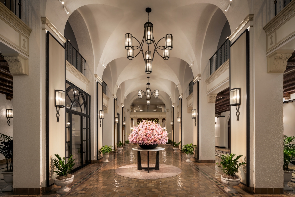 A grand resort hallway featuring vaulted white ceilings, wrought-iron chandeliers, tiled flooring, wall sconces, and a round center table displaying a large arrangement of pink flowers.